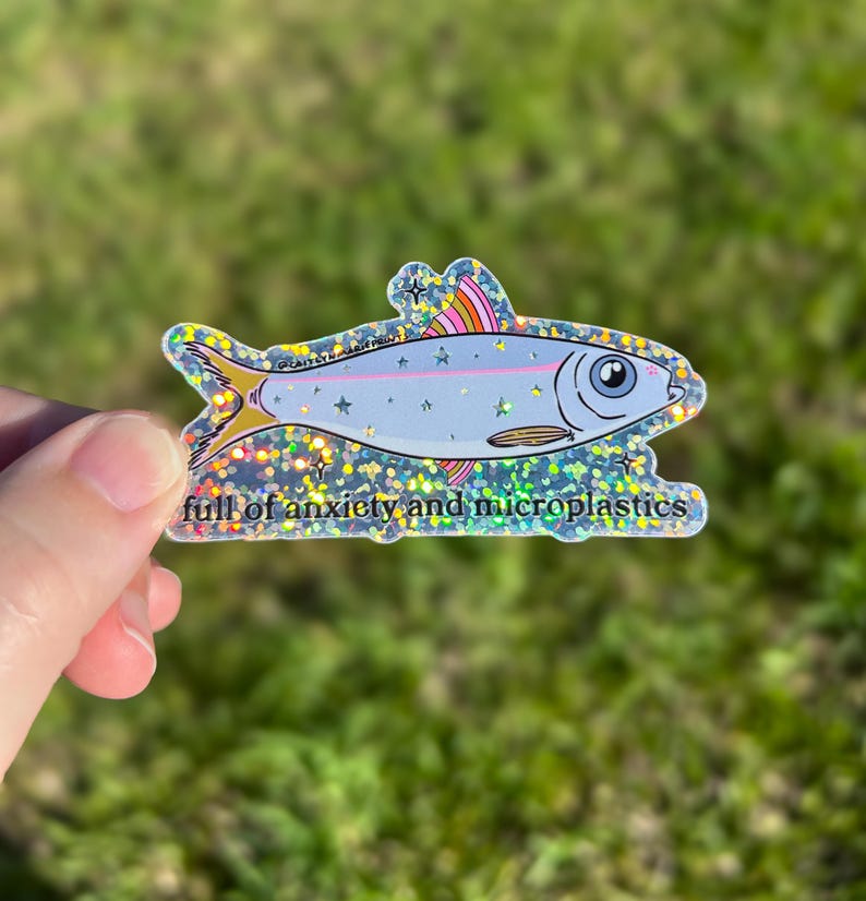 May include: A holographic sticker in the shape of a fish. The fish is light blue with a pink stripe and rainbow-colored fins. The text "full of anxiety and microplastics" is at the bottom of the sticker.