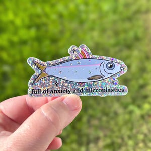 May include: A sticker featuring a stylized fish with a light blue body, gold tail, and rainbow-colored fins. The text "full of anxiety and microplastics" is below the fish. The sticker has a glittery, iridescent border.
