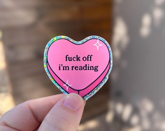 Può includere: Un adesivo a forma di cuore rosa con la scritta nera "fuck off i'm reading". L'adesivo ha un bordo olografico e due disegni a forma di stella bianca. L'adesivo è tenuto tra le dita.