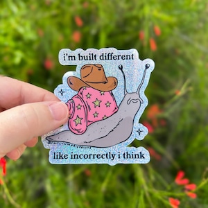 May include: A holographic sticker featuring a cartoon snail wearing a cowboy hat and a pink shell decorated with stars. The sticker has the text "i'm built different" and "like incorrectly i think".