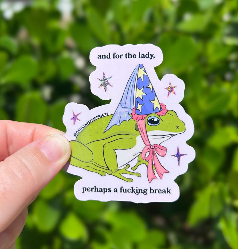 Pu&ograve; includere: Un adesivo a forma di rana verde con un cappello da mago con stelle blu e gialle e un fiocco rosa. L'adesivo ha il testo "and for the lady, perhaps a fucking break". L'adesivo &egrave; tenuto in mano.