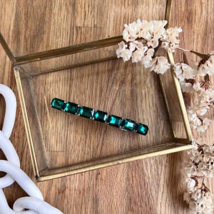 Puede incluir: Una pinza para el pelo de metal rectangular con piedras de imitación de esmeralda verde. La pinza es brillante.