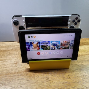 May include: A black Nintendo Switch console with a yellow stand. The screen displays the game "Pokémon: Let's Go, Eevee!" with other game icons visible. The console is connected to a white handheld gaming device.