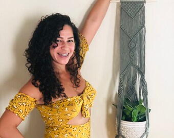 Pode incluir: Uma mulher vestindo um vestido amarelo floral está em frente a uma parede com um suporte de planta de macramê. O suporte é feito de fio cinza e tem um vaso de cerâmica branco com uma planta verde dentro.
