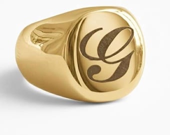 May include: A polished gold signet ring featuring a smooth, rounded face with a deeply engraved, dark-toned letter G in a cursive script font. The band has a thick, solid construction with a reflective, high-shine metallic finish throughout.