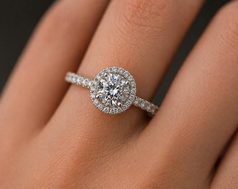 Könnte beinhalten: Ein silberfarbener Ring mit einem großen, runden, im Brillantschliff geschliffenen Mittelstein, der von einem Kranz aus kleineren, rund geschliffenen Steinen umgeben ist. Die Ringschiene ist schmal und mit einer Reihe winziger, funkelnder Steine besetzt.