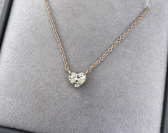 Può includere: Una collana delicata con un singolo ciondolo a forma di cuore incastonato in un metallo dai toni caldi. Il ciondolo è sospeso a una sottile catenina di metallo abbinata, creando un gioiello semplice e raffinato su una superficie grigia.