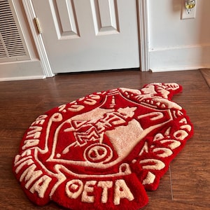 Puede incluir: Una alfombra roja y beige con un dise&ntilde;o de letras griegas. La alfombra tiene forma de escudo, con las letras griegas y otros s&iacute;mbolos en beige sobre un fondo rojo. La alfombra est&aacute; sobre un suelo de madera.