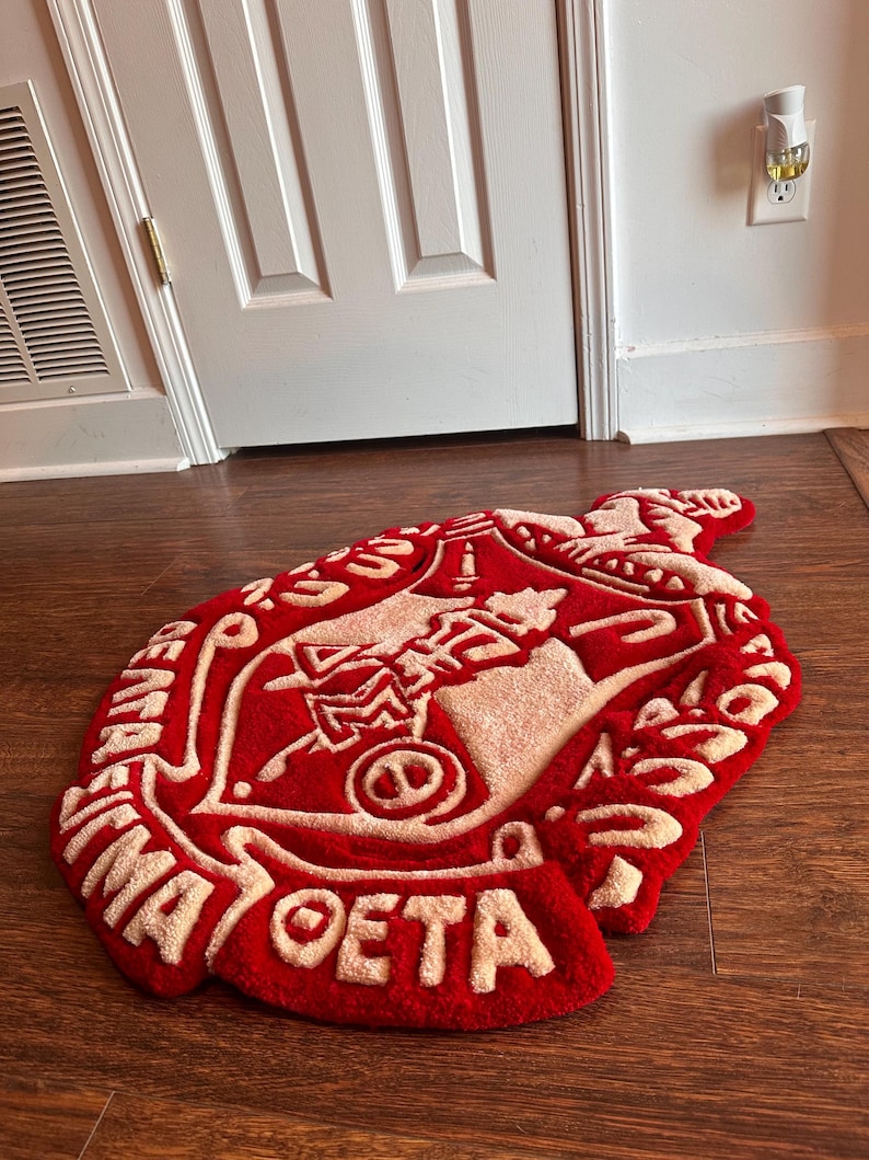 Puede incluir: Una alfombra roja y beige con un dise&ntilde;o de letras griegas. La alfombra tiene forma de escudo, con las letras griegas y otros s&iacute;mbolos en beige sobre un fondo rojo. La alfombra est&aacute; sobre un suelo de madera.