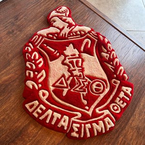 Puede incluir: Una alfombra roja y beige con forma de escudo, con letras griegas y un emblema central. La alfombra tiene un pelo grueso y texturizado y est&aacute; colocada sobre un suelo de madera. El dise&ntilde;o incluye las palabras "Delta Sigma Theta".