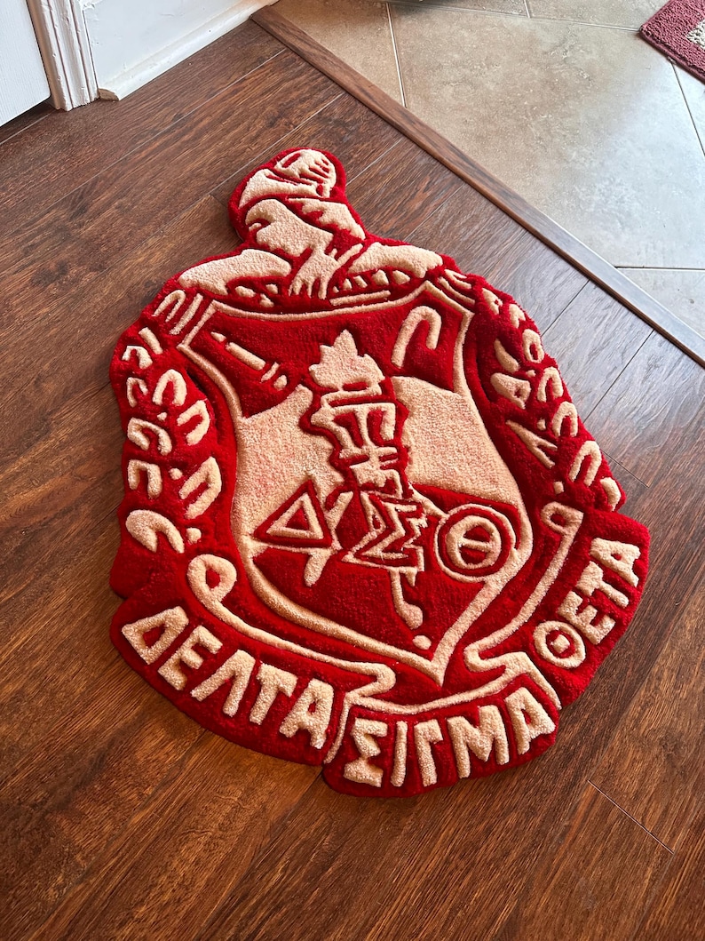 Puede incluir: Una alfombra roja y beige con forma de escudo, con letras griegas y un emblema central. La alfombra tiene un pelo grueso y texturizado y est&aacute; colocada sobre un suelo de madera. El dise&ntilde;o incluye las palabras "Delta Sigma Theta".