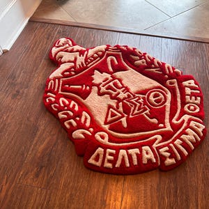 Puede incluir: Una alfombra roja y beige con un dise&ntilde;o de letras griegas. La alfombra tiene forma de escudo y presenta letras y s&iacute;mbolos griegos. La alfombra est&aacute; colocada sobre un suelo de madera.