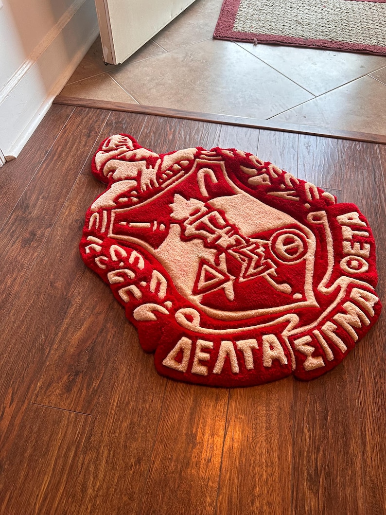 Puede incluir: Una alfombra roja y beige con un dise&ntilde;o de letras griegas. La alfombra tiene forma de escudo y presenta letras y s&iacute;mbolos griegos. La alfombra est&aacute; colocada sobre un suelo de madera.