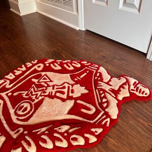 Puede incluir: Una alfombra roja y crema con un dise&ntilde;o abstracto &uacute;nico. La alfombra tiene una forma irregular y presenta un gr&aacute;fico central con texto en los bordes. La alfombra est&aacute; colocada sobre un suelo de madera.