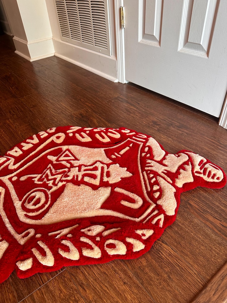 Puede incluir: Una alfombra roja y crema con un dise&ntilde;o abstracto &uacute;nico. La alfombra tiene una forma irregular y presenta un gr&aacute;fico central con texto en los bordes. La alfombra est&aacute; colocada sobre un suelo de madera.