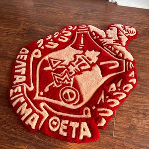 Puede incluir: Una alfombra roja y crema con un dise&ntilde;o de emblema detallado. La alfombra presenta un emblema central rodeado de texto, con una superficie texturizada y afelpada. La alfombra est&aacute; colocada sobre un suelo de madera.