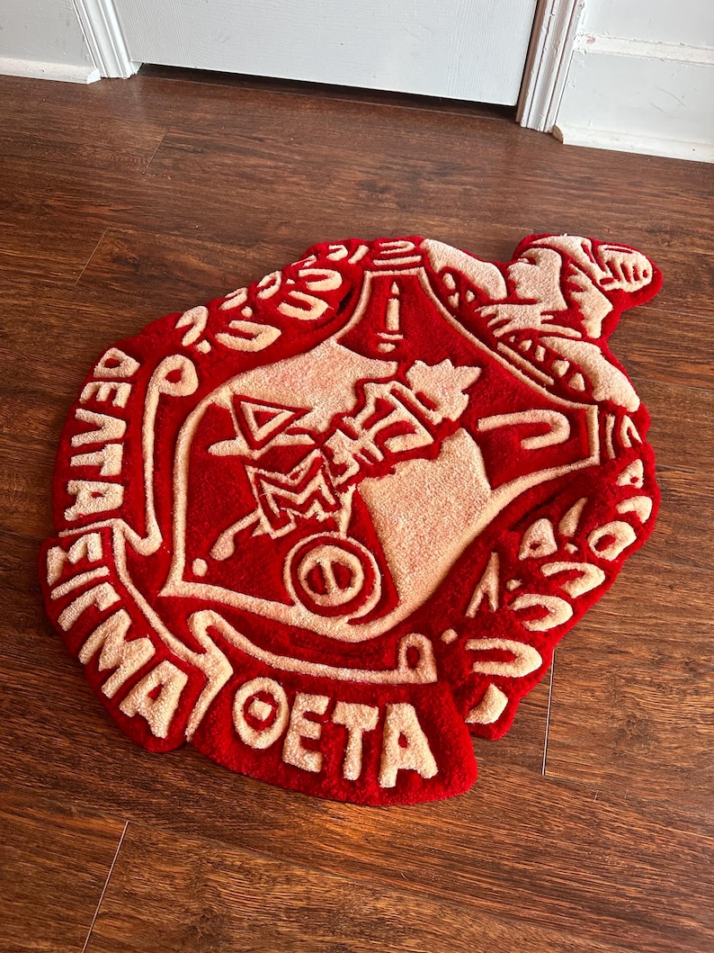 Puede incluir: Una alfombra roja y crema con un dise&ntilde;o de emblema detallado. La alfombra presenta un emblema central rodeado de texto, con una superficie texturizada y afelpada. La alfombra est&aacute; colocada sobre un suelo de madera.