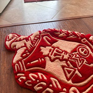 Puede incluir: Una alfombra roja y beige con una forma y dise&ntilde;o &uacute;nicos. La alfombra presenta un fondo rojo intenso con detalles beige, creando un contraste llamativo. El dise&ntilde;o incluye formas y patrones abstractos, agregando un toque moderno al revestimiento del suelo.