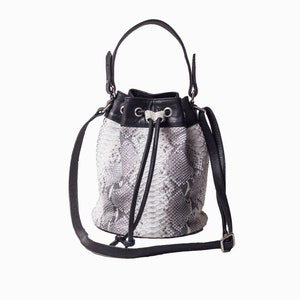 May include: A bucket bag with a snakeskin pattern in shades of gray and white. The bag features a black leather drawstring closure, a top handle, and a detachable shoulder strap. The bag has silver-toned hardware.