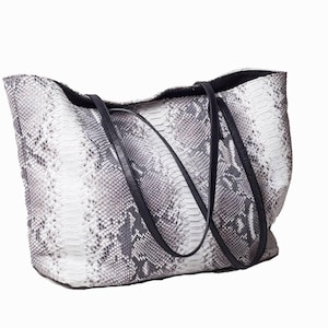 Könnte beinhalten: Eine große Tragetasche mit Schlangenmuster in Grau- und Weißtönen. Die Tasche hat schwarze Ledergriffe und ein schwarzes Innenfutter. Die Tasche ist zum Über-die-Schulter-Tragen konzipiert.