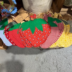 May include: A collection of handmade strawberry-shaped fabric coasters. The coasters feature red, white, and yellow patterns, including polka dots, gingham, and floral designs. Each coaster has a green felt leaf and a button detail.