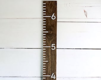 May include: A brown wooden ruler with white markings and silver numbers from 3 to 6. The ruler is mounted on a white wall.