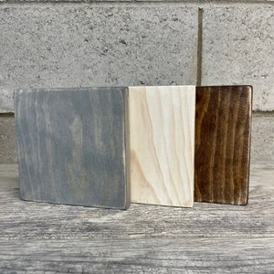 May include: Three wooden squares, one painted grey, one painted white, and one painted brown. The squares are arranged side by side on a wooden surface.