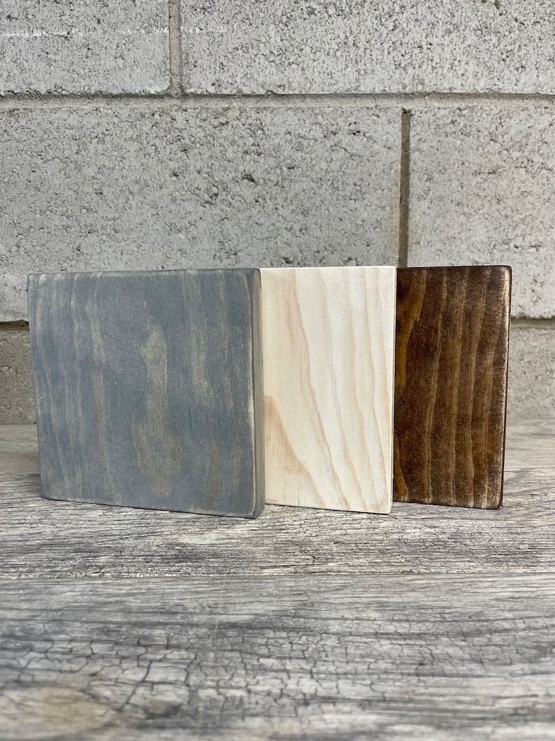 May include: Three wooden squares, one painted grey, one painted white, and one painted brown. The squares are arranged side by side on a wooden surface.