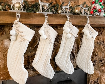 Peut inclure: Quatre chaussettes en tricot blanc avec des pompons accrochés à une cheminée en bois. Chaque chaussette a une étiquette de nom en bois avec les noms "Papa", "Maman", "La Fille" et "Jackson".