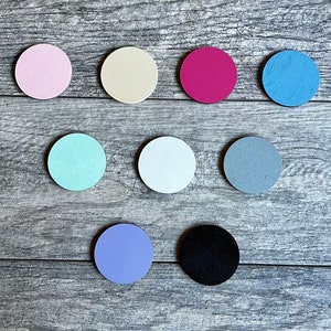 Puede incluir: Diez discos redondos de madera en varios colores, incluyendo rosa, beige, rojo, azul, azul claro, blanco, gris, morado y negro. Los discos est&aacute;n dispuestos en semic&iacute;rculo sobre una superficie de madera.