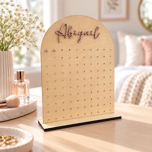 May include: A light-colored wooden earring display stand with an arched top. The stand features a grid of small holes for organizing jewelry and is personalized with the name Abigail in raised script lettering attached to the upper center.