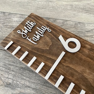 May include: A brown wooden growth chart with white painted lines and a silver number "6" at the top. The words "Smith Family" are written in silver cursive lettering at the top of the chart.