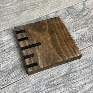 May include: A square wooden coaster with a dark brown stain and black painted lines across the top.