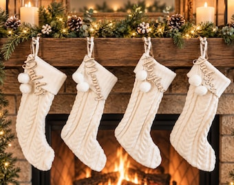 May include: Four cream-colored cable knit Christmas stockings hanging in a row. Each stocking features a ribbed cuff, two white pom-poms, and a wooden name tag with the script text Daddy, Mommy, Haylen, or Jackson attached to the hanging loop.