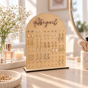 May include: A light-colored wooden earring display stand with an arched top featuring the name Abigail in raised script lettering. The stand has a grid of small holes for organizing various gold-toned earrings, including studs, hoops, and dangling styles.