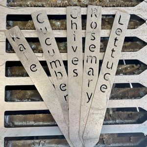 May include: Five metal plant markers with the words Kale, Cucumber, Chives, Rosemary, and Lettuce written on them.