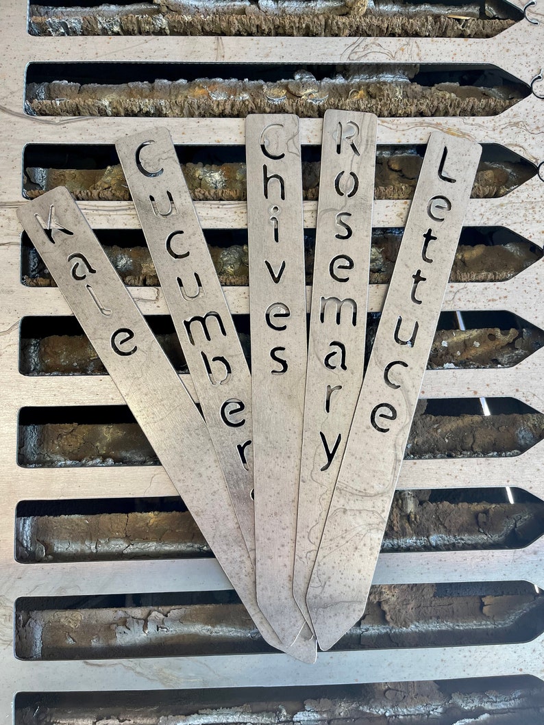 May include: Five metal plant markers with the words Kale, Cucumber, Chives, Rosemary, and Lettuce written on them.