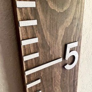 May include: A wooden growth chart with white painted markings and a silver number "5" on a brown wooden background.