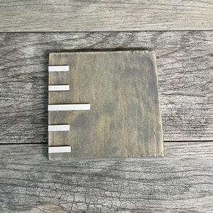 May include: A square wooden coaster with a grey stain and five silver metal strips on the left side.