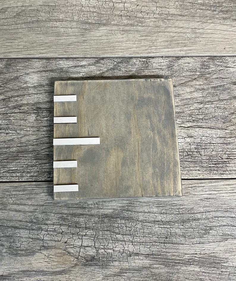 May include: A square wooden coaster with a grey stain and five silver metal strips on the left side.