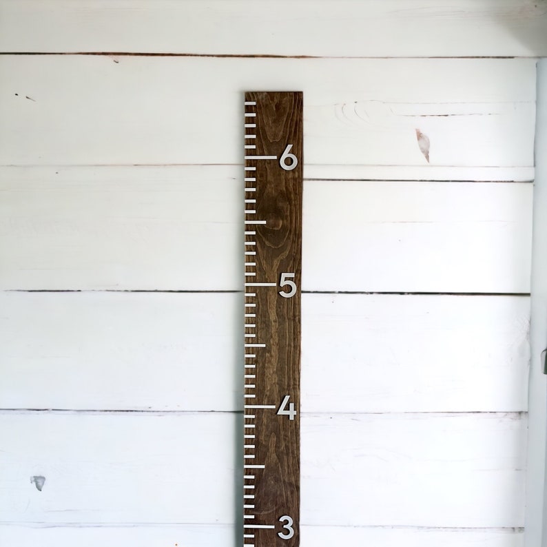 May include: A wooden growth chart with white markings and silver numbers from 3 to 6. The chart is mounted on a white wall.
