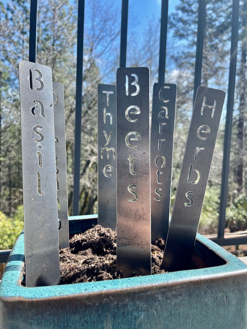 May include: Metal plant markers with the words "Basil", "Beets", "Carrots", "Herbs", and "Thyme" written on them. The markers are inserted into a teal ceramic planter filled with brown soil.