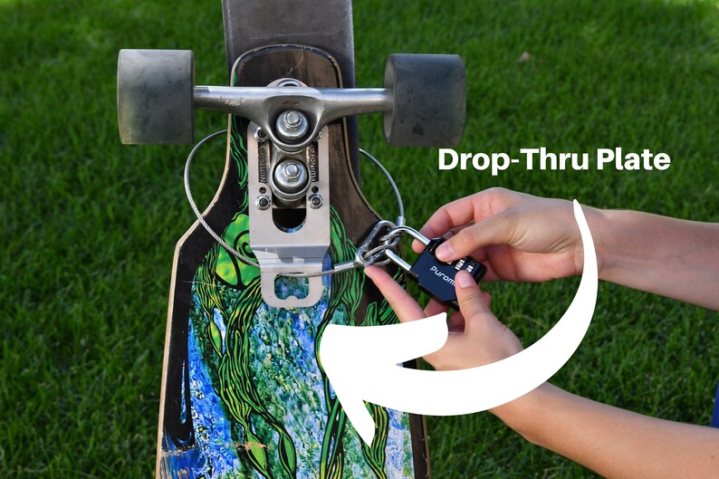 Puede incluir: Un candado de combinaci&oacute;n negro est&aacute; unido a un cable plateado que se enrosca a trav&eacute;s de una placa de metal en una tabla de skate larga. La placa est&aacute; etiquetada como "Drop-Thru Plate".