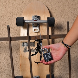 Puede incluir: Un candado de combinaci&oacute;n negro con un cable plateado unido a una tabla de skate de madera. La tabla de skate est&aacute; asegurada a una valla met&aacute;lica con el cable. La tabla de skate tiene un gr&aacute;fico en blanco y negro de una persona haciendo skate con el texto "Skate Snatch Crew" en ella.