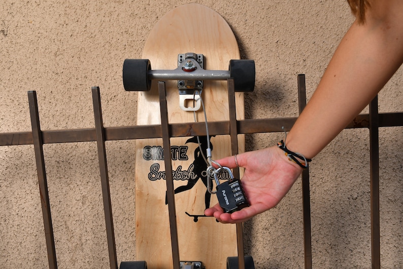 Puede incluir: Un candado de combinaci&oacute;n negro con un cable plateado unido a una tabla de skate de madera. La tabla de skate est&aacute; asegurada a una valla met&aacute;lica con el cable. La tabla de skate tiene un gr&aacute;fico en blanco y negro de una persona haciendo skate con el texto "Skate Snatch Crew" en ella.