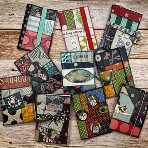 Könnte beinhalten: Eine Sammlung handgefertigter Notizbuchhüllen im Patchwork-Stil in verschiedenen Farben und Mustern. Jede Hülle weist einzigartige Stoffkombinationen auf, darunter Blumen-, geometrische und Tiermuster. Einige Hüllen haben elastische Verschlüsse oder dekorative Akzente.
