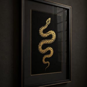 May include: A framed wall art piece featuring a gold snake illustration on a solid black background. The artwork is encased in a thin black frame with gold inner detailing, presented in a vertical orientation with a clean, sharp aesthetic.