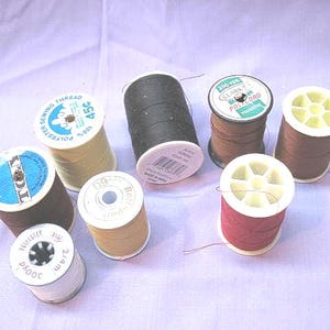 May include: A collection of spools of thread in various colors, including brown, cream, and red. The spools have white or cream-colored bases and tops, with labels displaying text and logos. The thread is likely polyester and suitable for sewing projects.