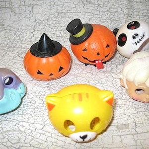 May include: A collection of Halloween-themed toys, including orange pumpkins with hats, a white skull, a yellow cat, and a blue and purple creature. The toys are made of plastic and have various facial expressions.