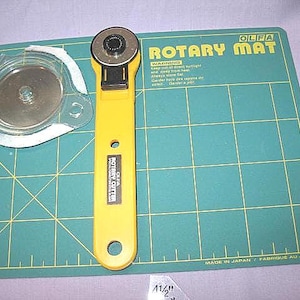 May include: A yellow Olfa rotary cutter with a black blade and a green Olfa rotary mat. The mat has grid lines and the text "Rotary Mat". A clear plastic blade cover is also present.